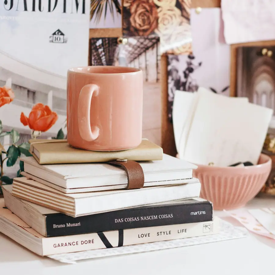 A pink mug resting on a stack of books and notebooks, beside roses on a bright desk